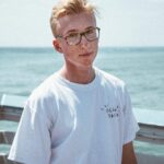 Young man in glasses wearing a white shirt, standing by the sea on a sunny day.