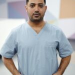 Portrait of a confident male nurse standing, wearing a light blue scrub suit in a modern medical setting.