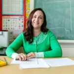 Engaging image of a smiling teacher seated at a desk in a classroom environment.